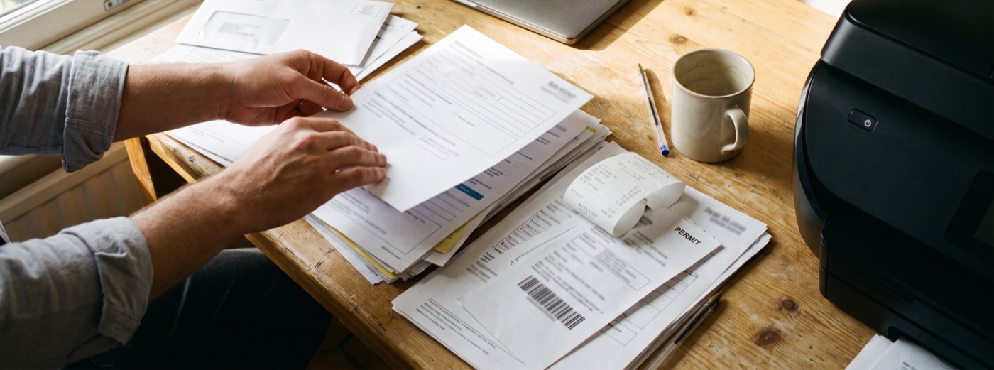 Hands sorting current business paperwork on a desk — printed forms, a windowed envelope, a thermal receipt, a barcoded permit